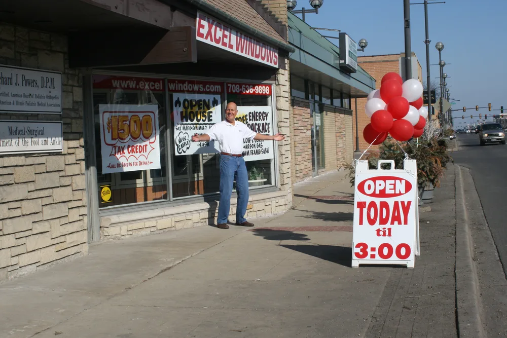 An Excel open house — a team member welcoming visitors out front by the OPEN TODAY sign and balloons.