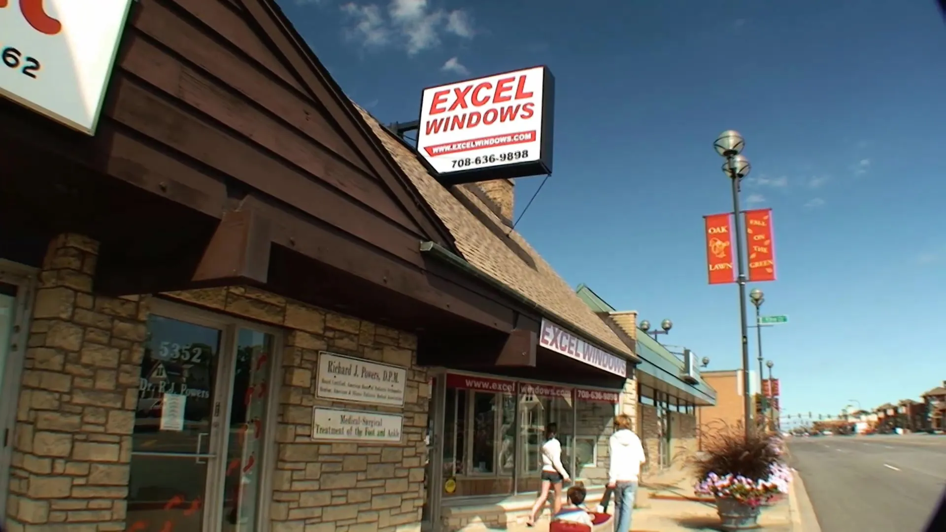 Excel's Oak Lawn storefront in summer, the EXCEL WINDOWS sign up on the roof and a family on the sidewalk.